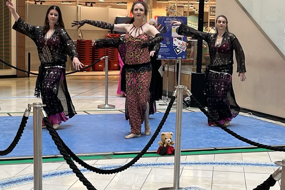 Drei Bauchtänzerinnen in dunklen Kostümen auf einer Bühne, einen Arm nach links ausgestreckt.