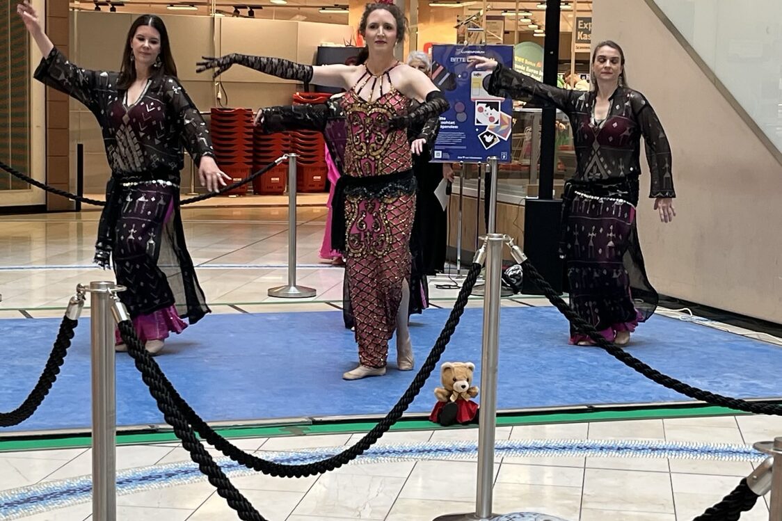 Drei Bauchtänzerinnen in dunklen Kostümen auf einer Bühne, einen Arm nach links ausgestreckt.