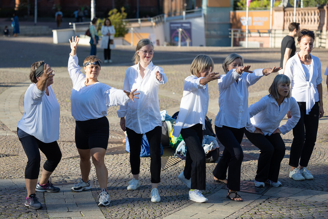 Eine Gruppe Frauen in weißen Hemden und schwarzen Hosen steht in einer Reihe und streckt die Arme nach vorn.