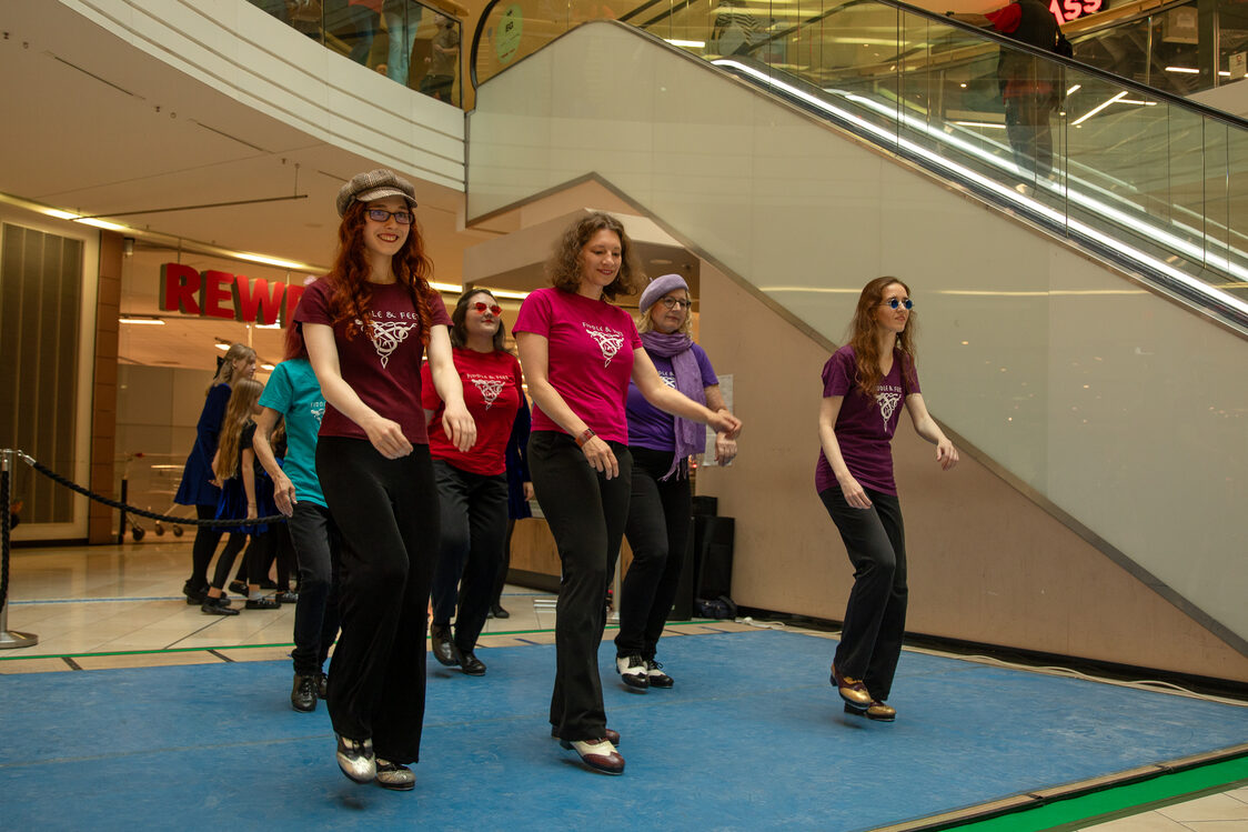 Sechs Frauen mit schwarzen Hosen und bunten T-Shirts tanzen in einem Einkaufszentrum.