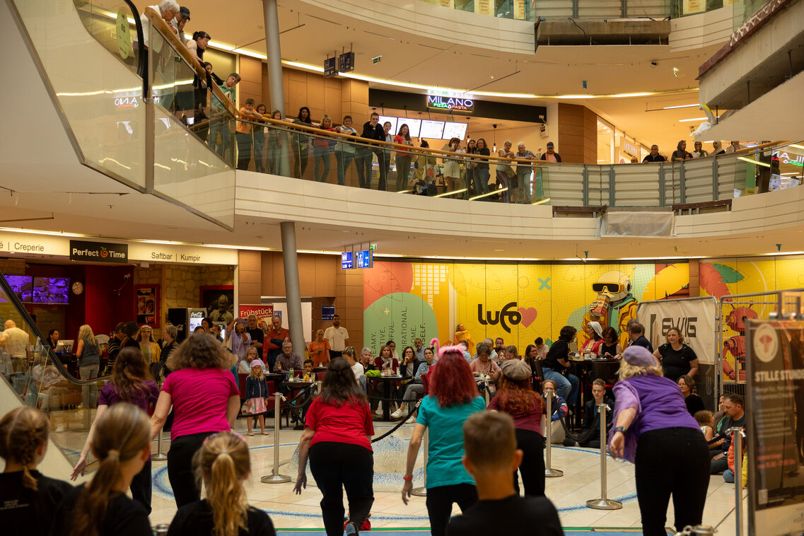 Sechs Frauen mit schwarzen Hosen und bunten T-Shirts tanzen in einem Einkaufszentrum. Menschen schauen zu.